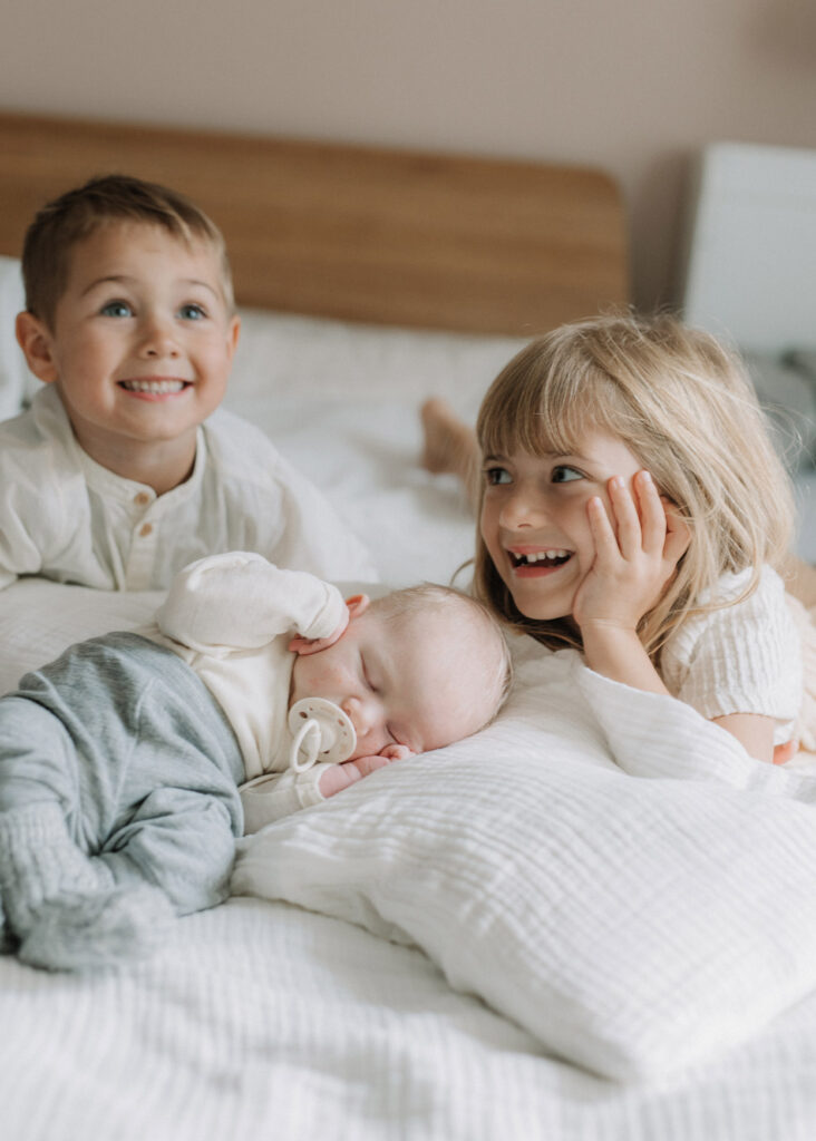 Geschwister sitzen mit neugeborenen Baby lachend auf dem Bett bei einen Neugeborenen-Fotoshooting in Berghaupten.